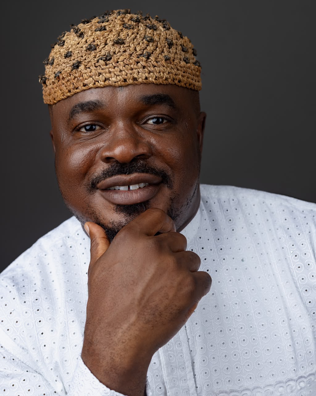 John Ifere smiling in white embroidered traditional attire and woven cap